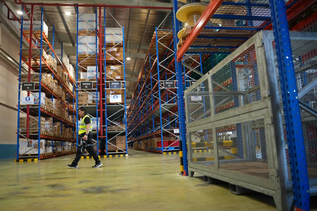 A worker in a yellow safety vest walks through a spacious warehouse with tall blue and orange shelves full of neatly stacked boxes, conveying efficiency.