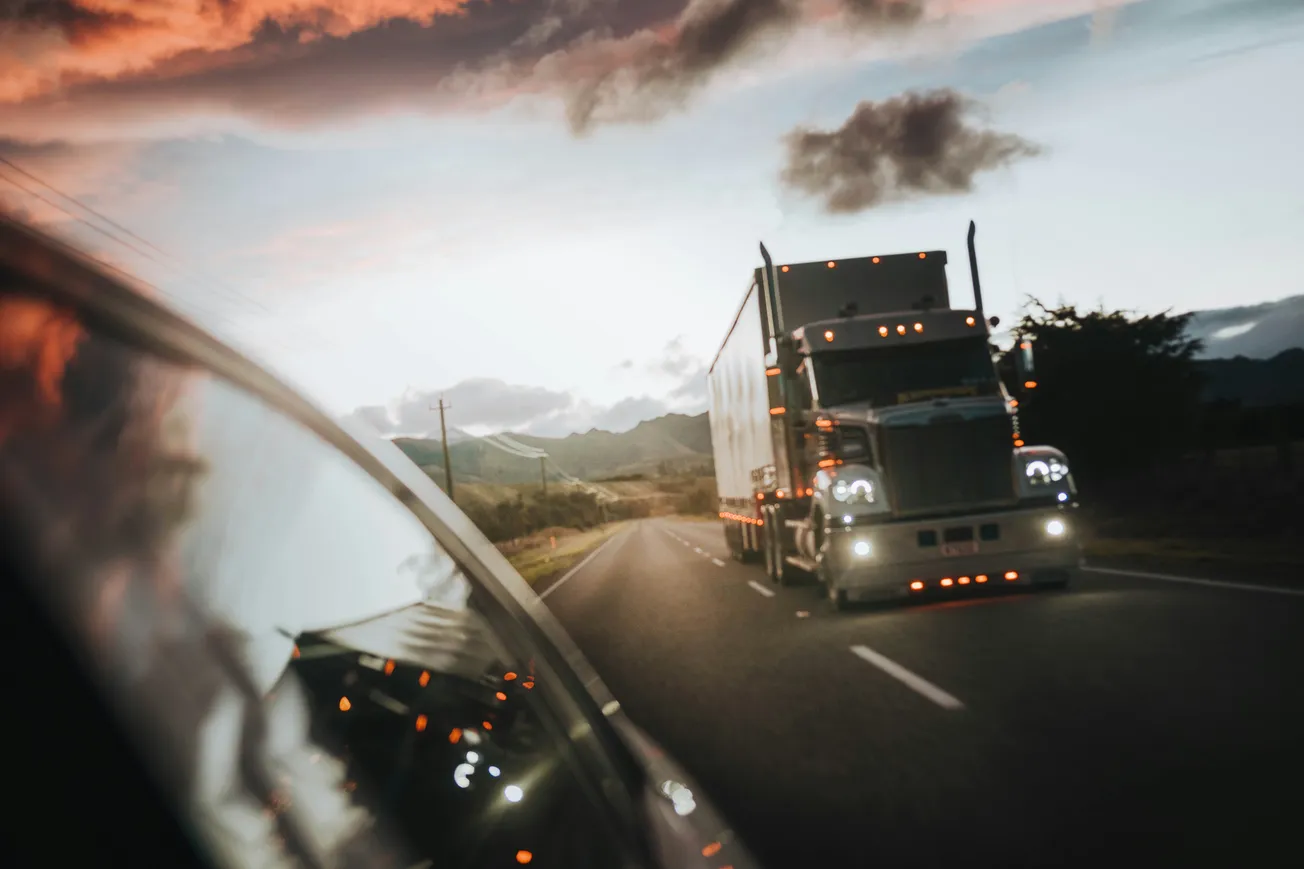 A large truck with shining headlights drives on an open road at sunset, reflected in a passing car's side mirror, with mountains and clouds in the background.