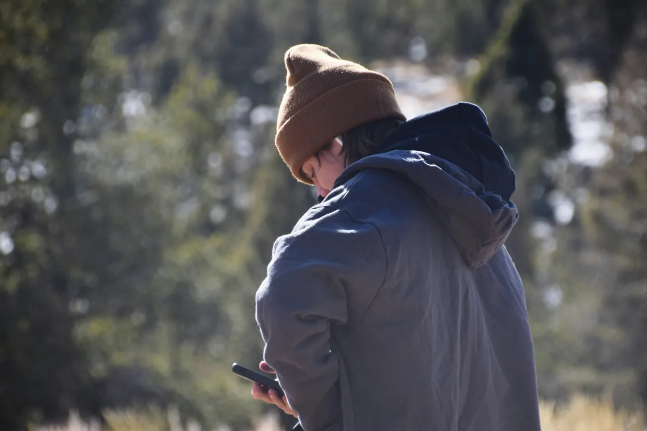 A person in a brown beanie and blue jacket looks at a smartphone in a forest. The scene conveys a sense of solitude and connection with nature.