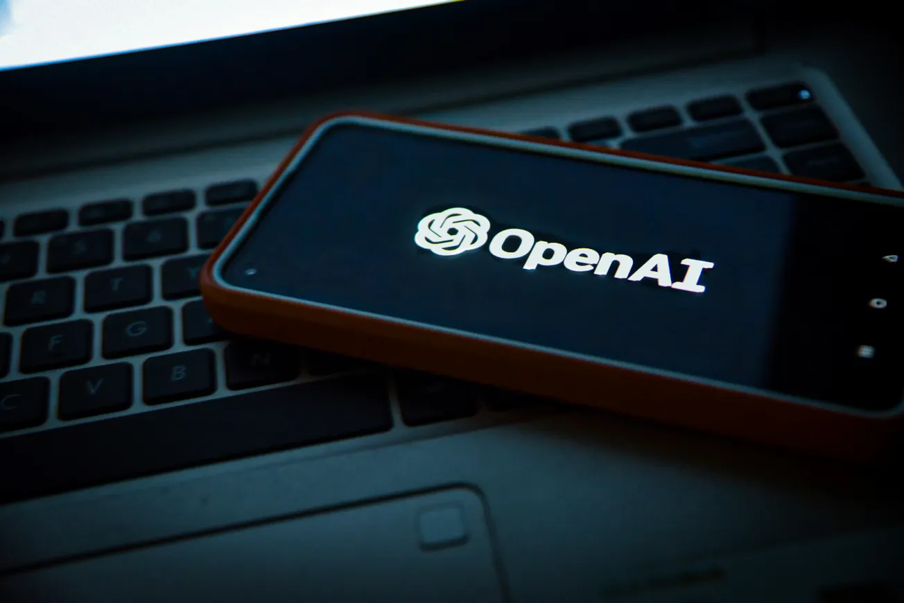 A smartphone displaying the OpenAI logo rests on a laptop keyboard. The image conveys a modern, tech-focused atmosphere, highlighting AI technology.