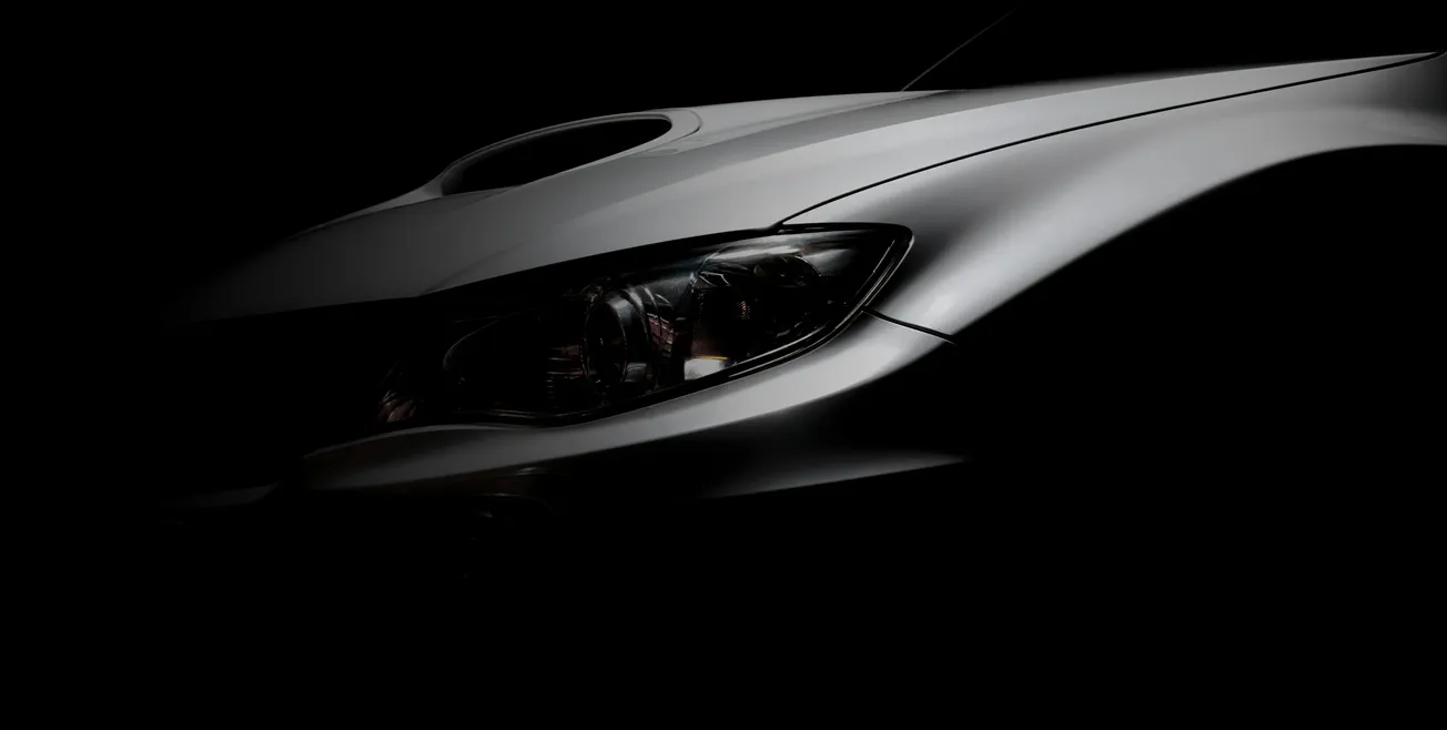 Close-up of a sleek car front in shadow, showing its glossy silver hood and sharp headlight. The lighting adds a mysterious, sophisticated tone.