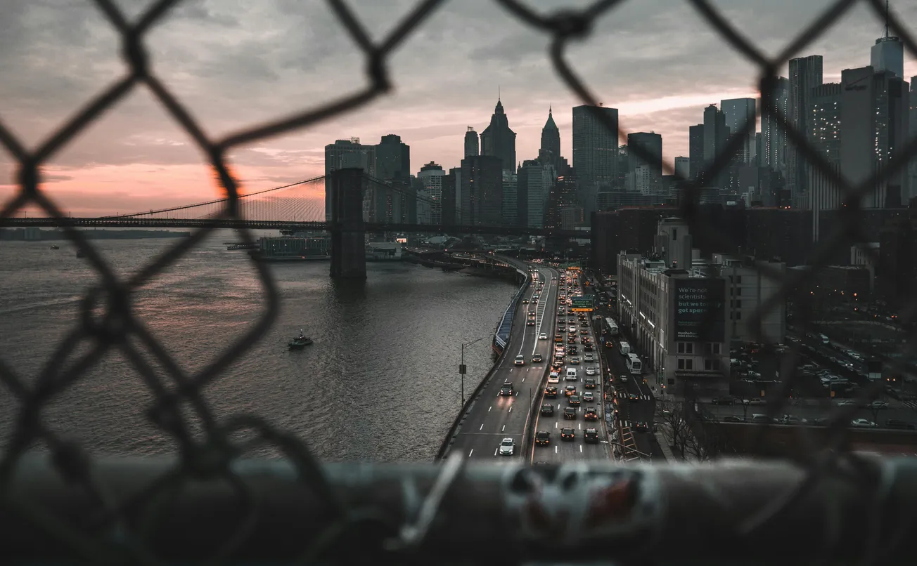 View of the city skyline at dusk through a chain-link fence. A bridge spans the river, and traffic moves along the road, creating a moody urban scene.