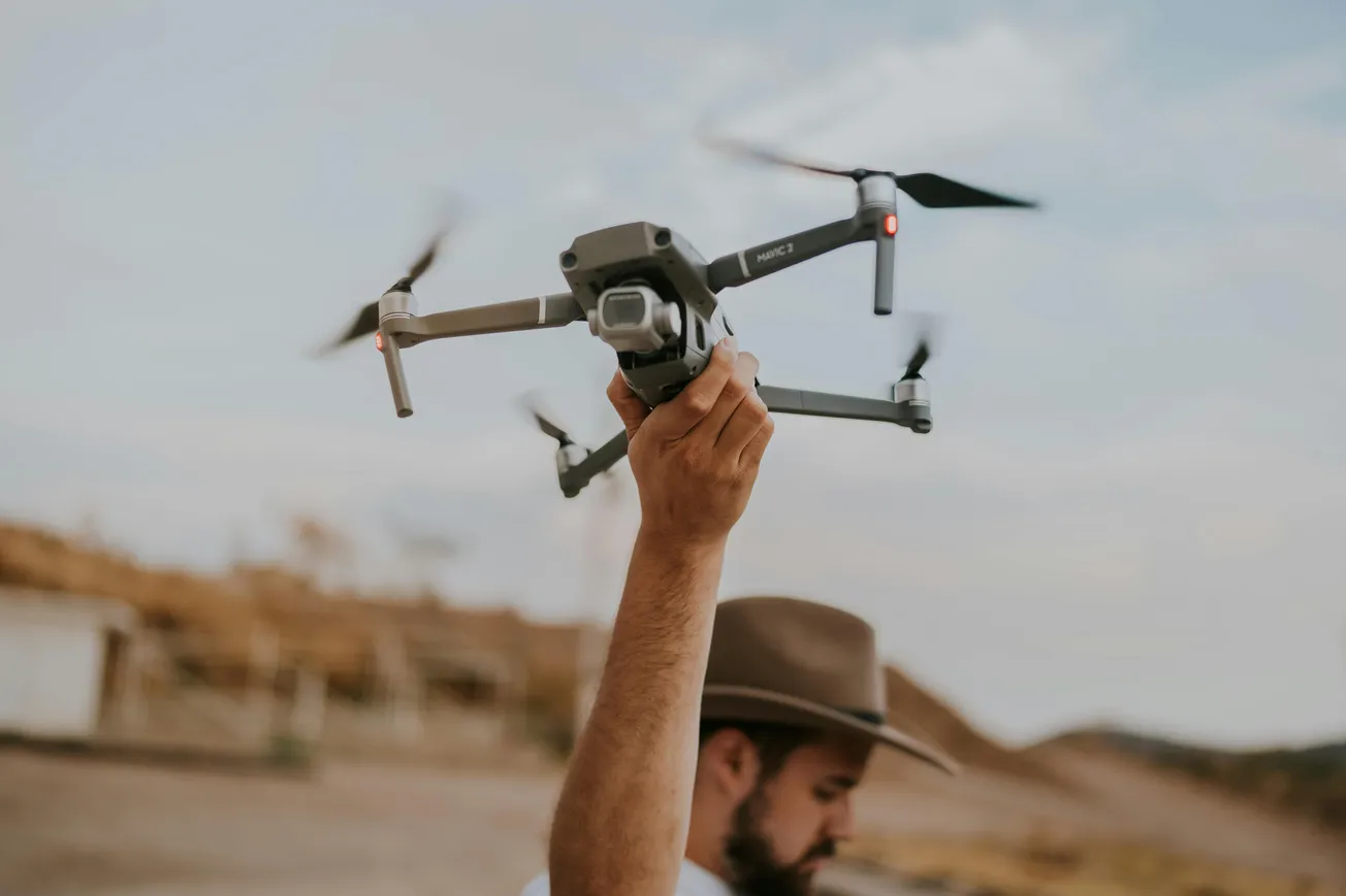 A person in a hat holds a drone mid-flight against a blurred outdoor landscape, conveying a sense of adventure and technology in nature.