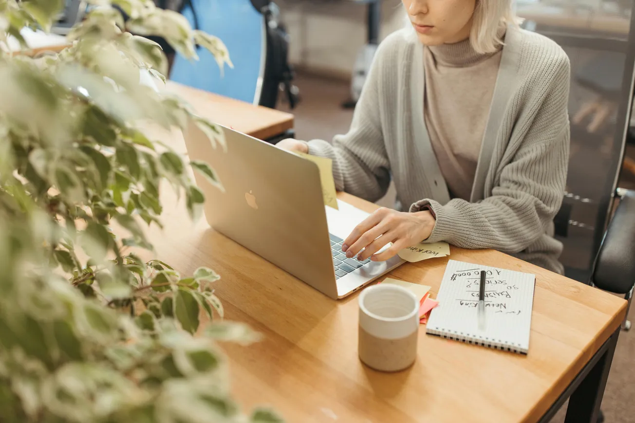 A person is working at a wooden desk with a laptop, surrounded by sticky notes and greenery. A notepad and a coffee cup add a calm, focused tone.