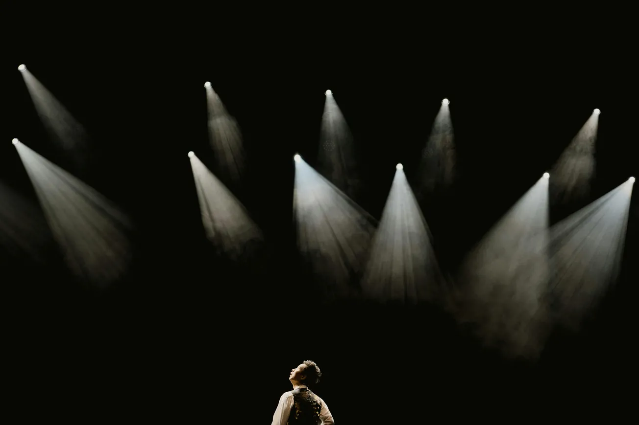 A person stands on stage, looking up at bright, dramatic spotlights in a dark theater. The scene conveys anticipation and focus.