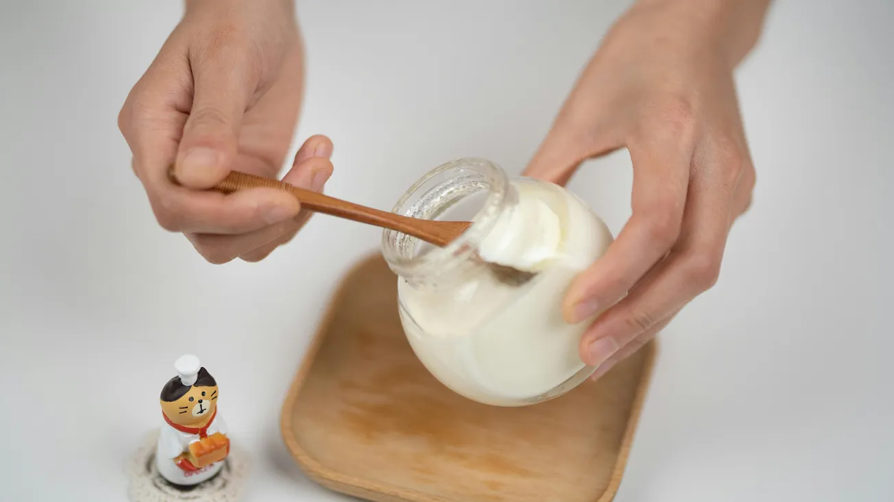 Hands holding a wooden spoon and a jar of creamy yogurt over a wooden tray. A small figurine of a chef with a cat-like face stands nearby, adding a whimsical touch.