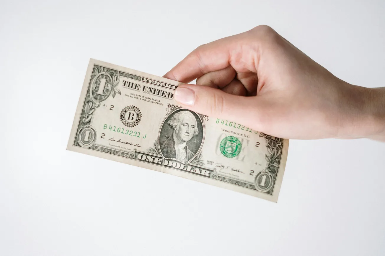 A hand holds a one-dollar bill against a plain white background. The bill features George Washington's portrait. The scene is minimal and focused.