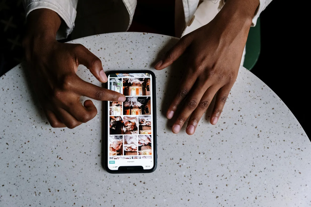 A person browses food photos on a smartphone placed on a round speckled table. One hand scrolls the screen, the other rests beside it, creating a focused mood.