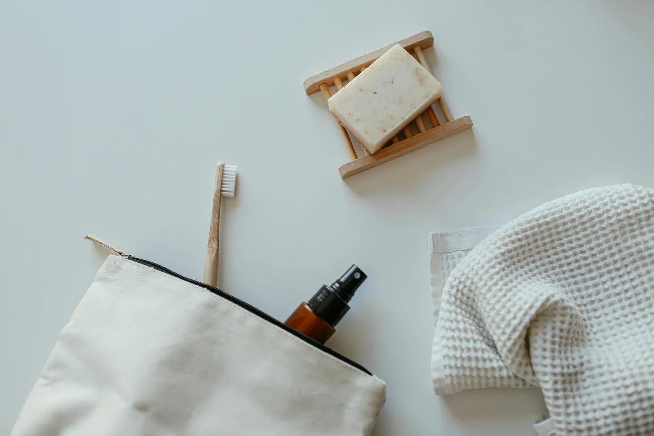 Top view of eco-friendly bathroom items on a white surface, including a bamboo toothbrush, soap on a wooden tray, spray bottle, and beige waffle towel.