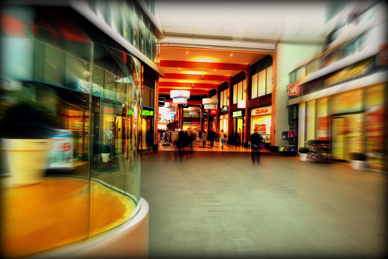 Blurred view of a bustling indoor shopping mall with bright lights, colorful storefronts, and people walking. The scene feels vibrant and lively.