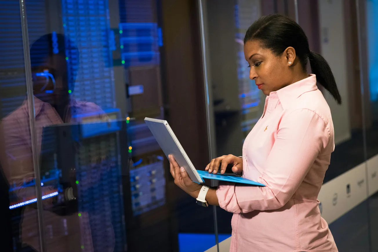 A woman in a pink shirt works on a laptop in a server room. She is focused, with the server lights reflecting blue on glass, creating a tech-centric atmosphere.