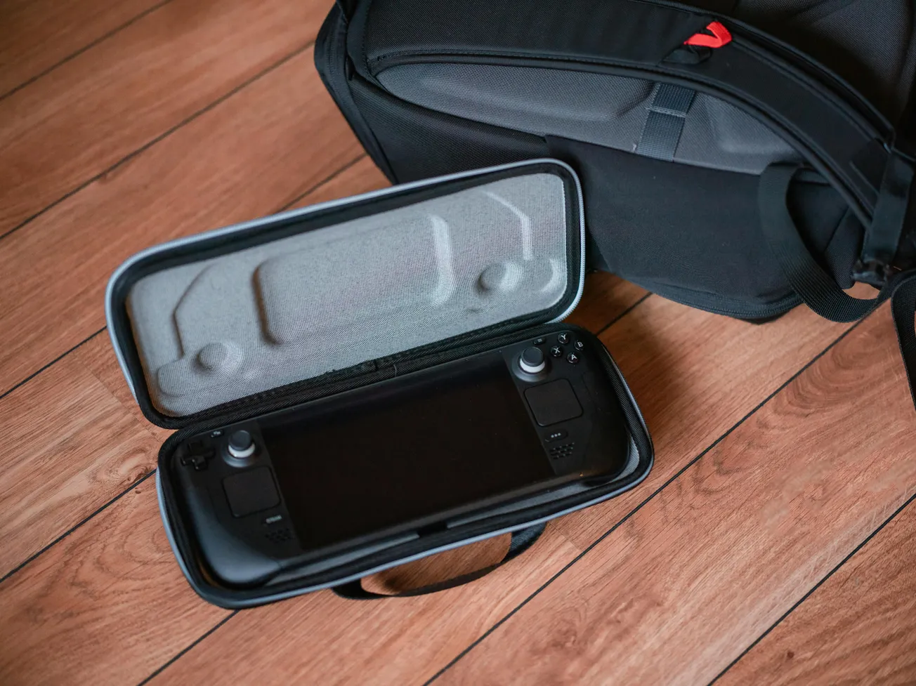 A gaming handheld device in an open case rests on a wooden floor. A black backpack is nearby. The scene conveys readiness for travel or gaming on the go.