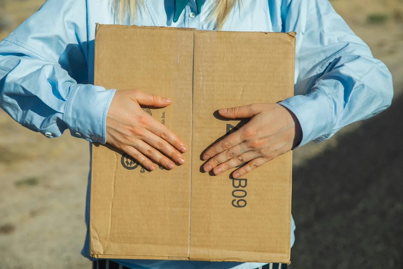 Person in a blue shirt holding a large cardboard box outdoors. The box is centered in the image, and the scene conveys a sense of activity and movement.