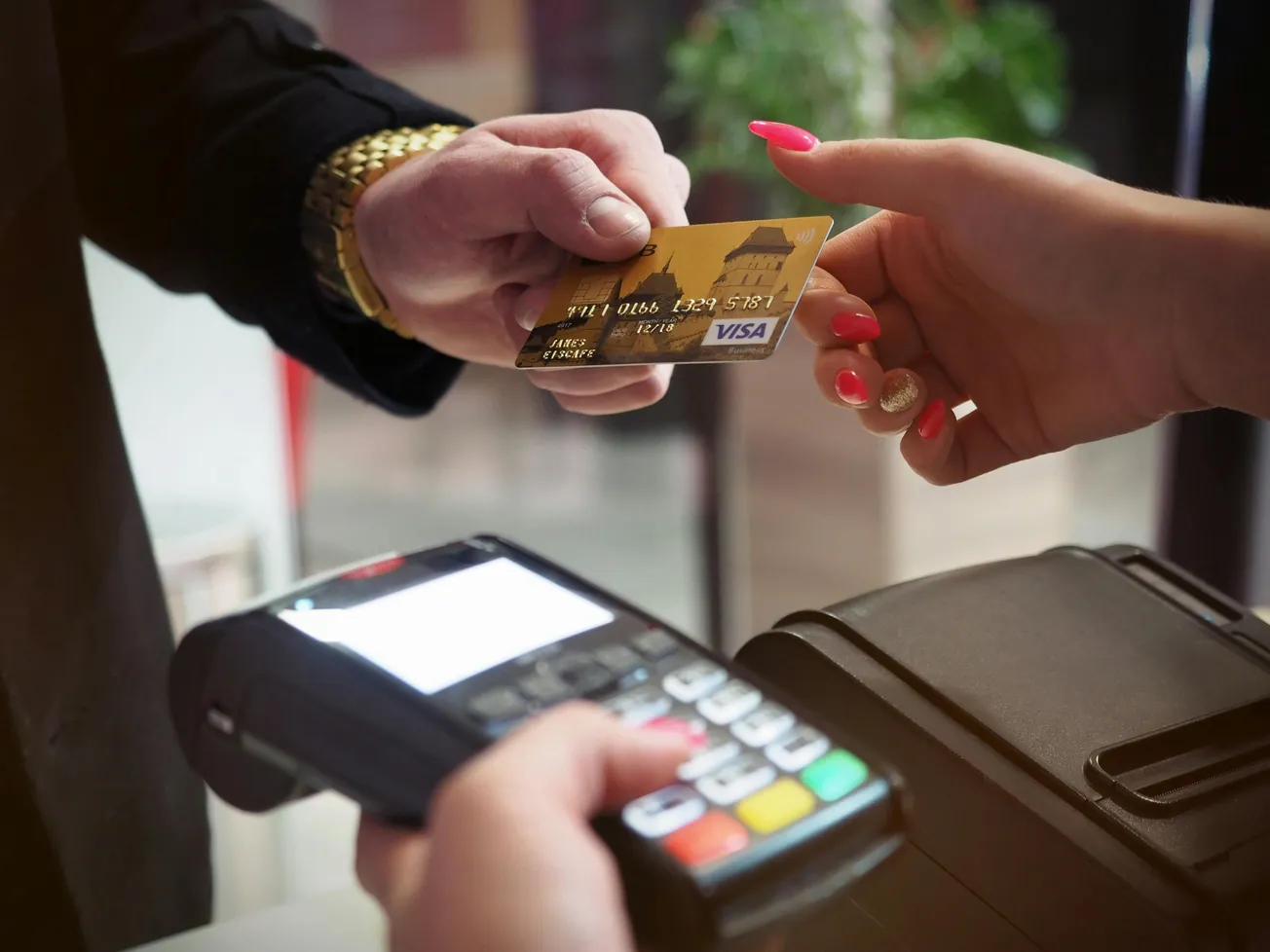 A person with red nails hands a Visa credit card to another with a gold watch. A payment terminal is held nearby, ready for transaction.