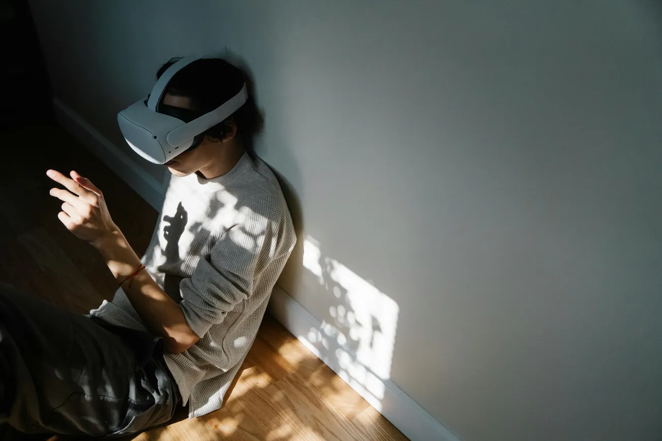 A person wearing a VR headset sits against a sunlit wall, immersed in virtual reality. Dappled light creates a serene, contemplative atmosphere.