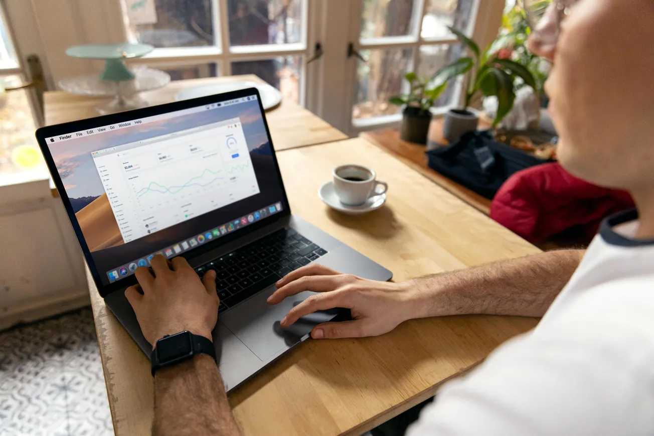A person at a wooden table works on a laptop displaying graphs. A cup of coffee and plants are nearby, creating a focused, home-office atmosphere.