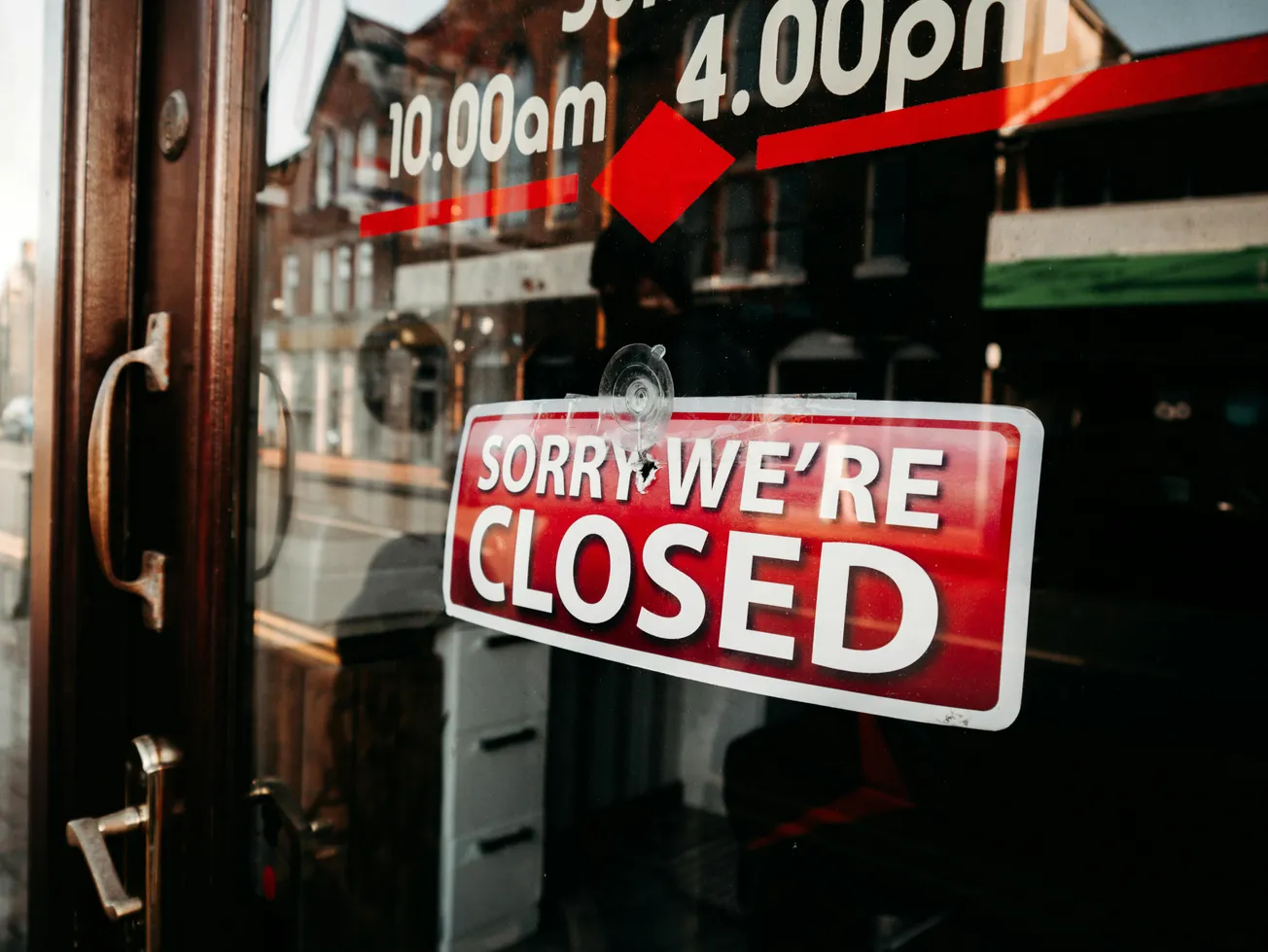 Storefront glass door with a red "Sorry, We're Closed" sign. Reflections show buildings and a street.