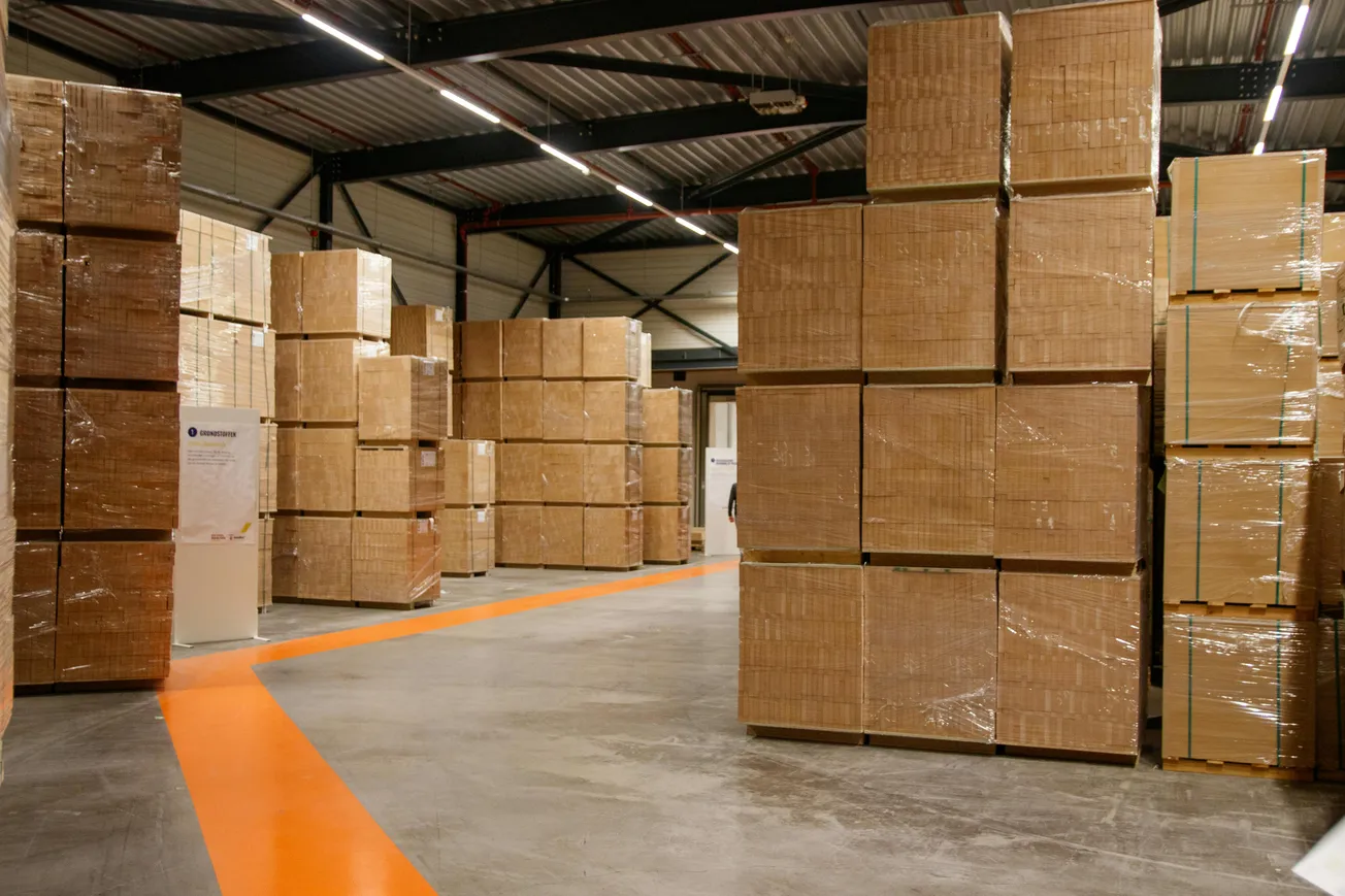 Spacious warehouse interior with tall stacks of sealed cardboard boxes on pallets, gray concrete floor, bright orange line, and high, industrial ceiling.