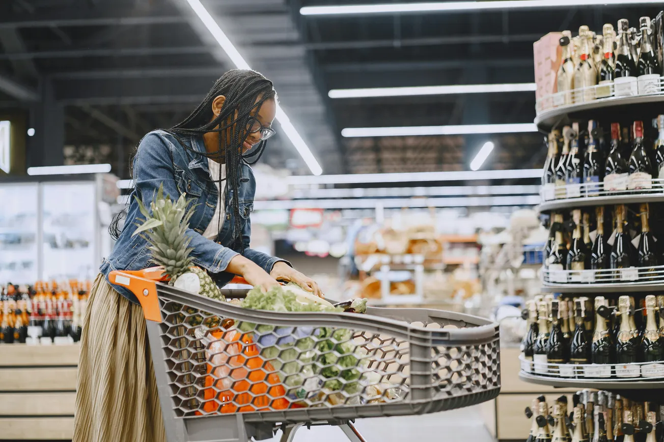 A woman in a denim jacket shops in a supermarket, pushing a cart full of produce, including pineapples and oranges, with wine bottles on shelves nearby.