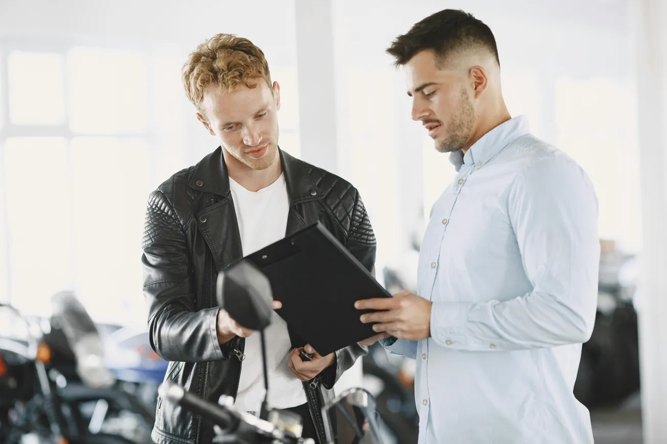 Two men stand in a motorcycle showroom. One in a leather jacket examines a clipboard held by the other in a light blue shirt, discussing details.