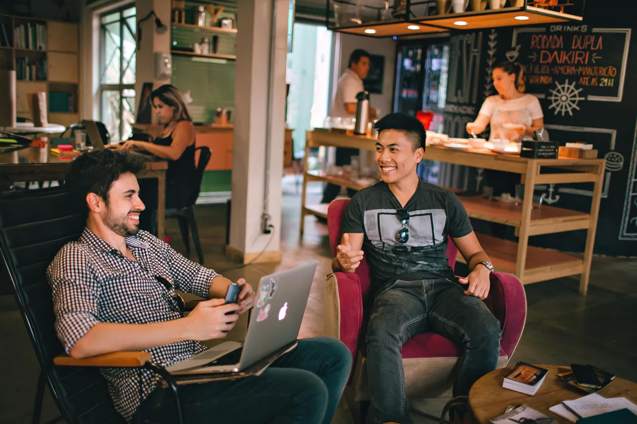 Two men engage in lively conversation at a café, each seated with a laptop. In the background, two people work at a counter, creating a relaxed and social atmosphere.