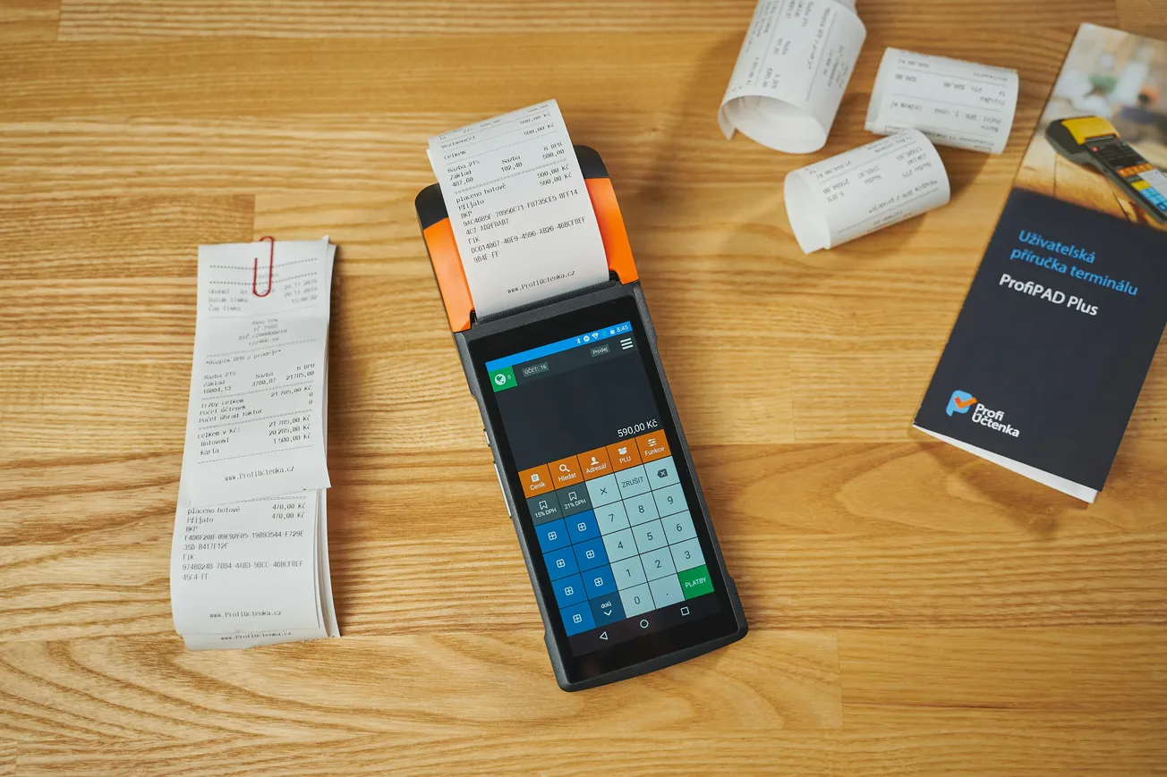 A payment terminal with a printed receipt sits on a wooden surface, surrounded by rolled receipts and a user manual. The scene conveys an organized, business-like atmosphere.
