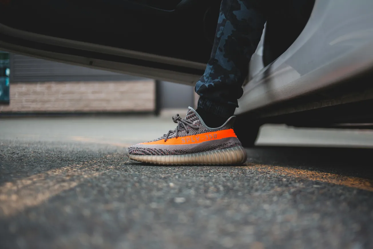 A person wearing gray and orange sneakers steps out of a car. The ground is asphalt, and the setting suggests a casual, urban vibe.