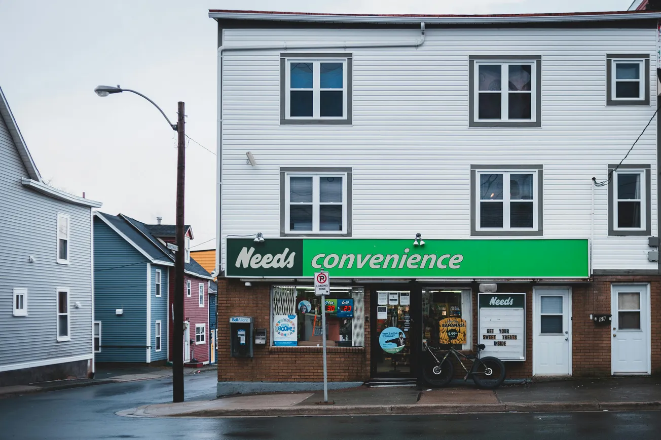 Facade of a small convenience store under a residential building. The sign reads "Needs Convenience" in green. A bicycle is parked by the entrance.
