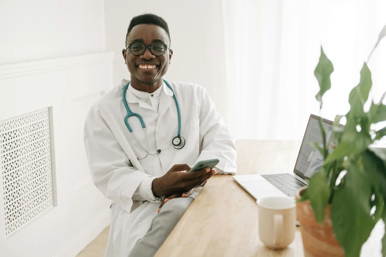 A smiling doctor wearing a white coat and stethoscope sits at a wooden table with a laptop and smartphone. A potted plant and mug are nearby, conveying a warm, professional atmosphere.
