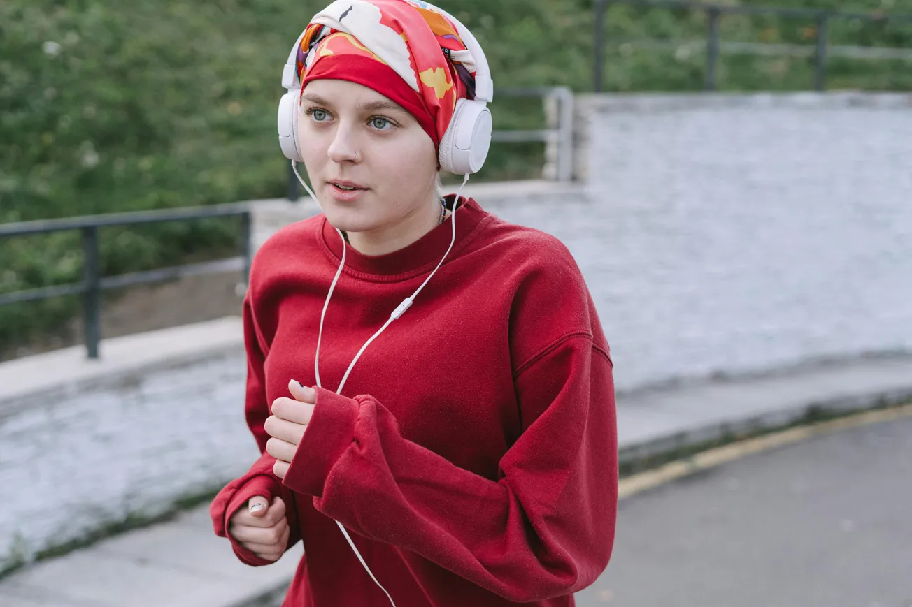 Young woman jogging outdoors, wearing a red sweater, colorful headscarf, and headphones. She appears focused and determined. Background: greenery and pathway.