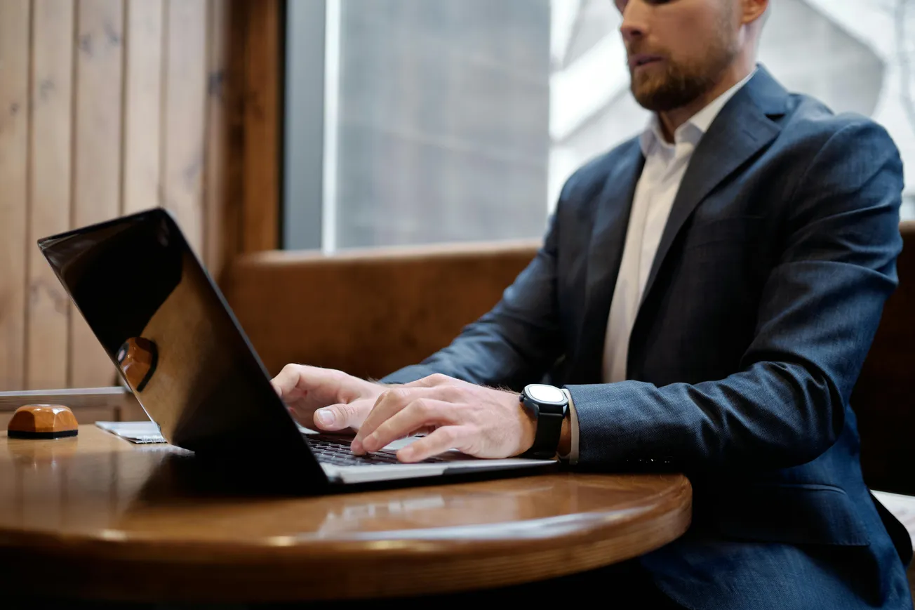 A man in a suit types on a laptop at a wooden table, conveying focus and professionalism. Natural light filters through a window, creating a calm atmosphere.