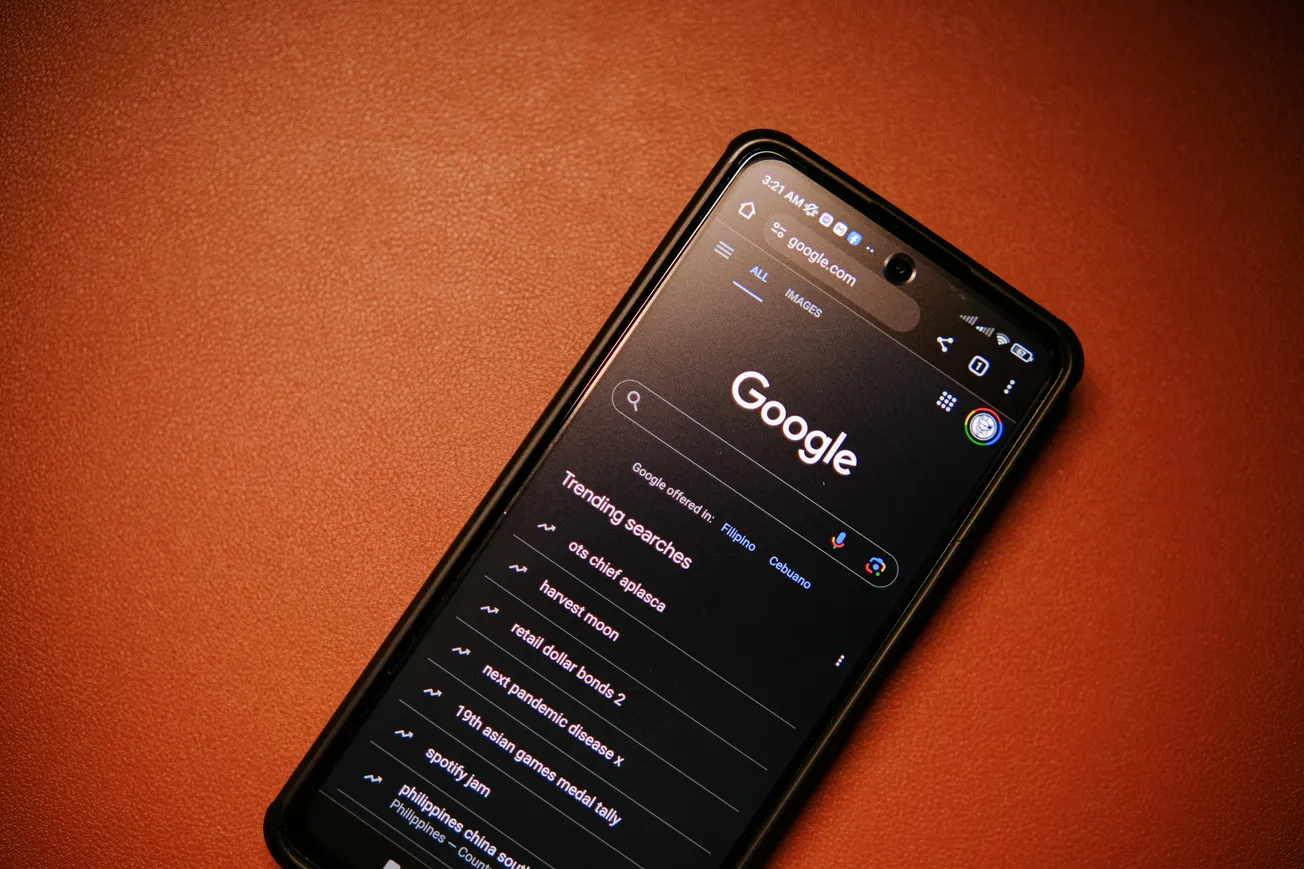 Smartphone on an orange surface displaying Google's dark mode search page with trending searches. The screen is lit, creating a focused and modern feel.