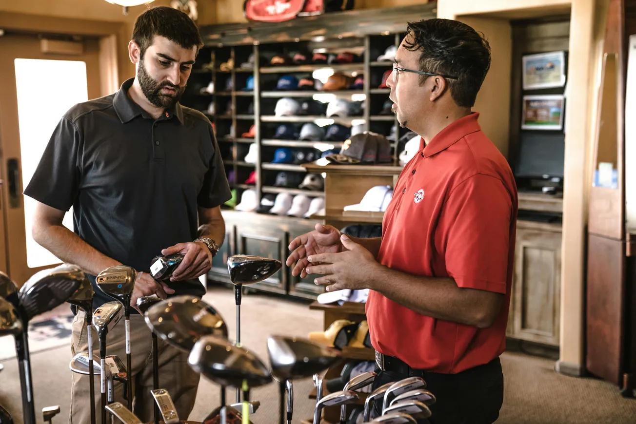 Two men are in a golf shop discussing golf clubs. The man on the left holds a club, listening intently, while the other gestures, explaining. Shelves with hats are in the background.