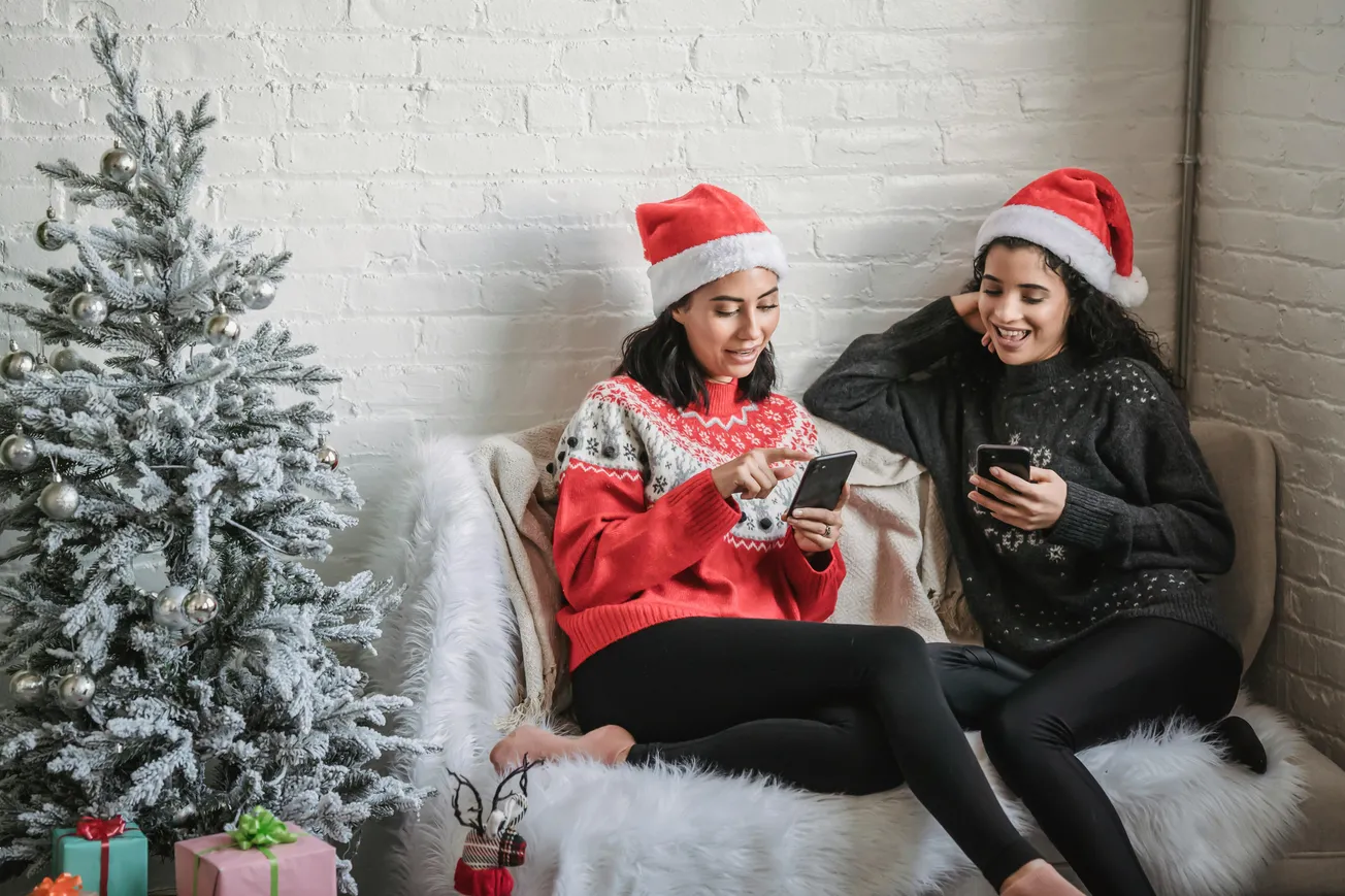 Two women wearing Santa hats and festive sweaters sit on a cozy couch, smiling while looking at their phones. A decorated Christmas tree with gifts is beside them.