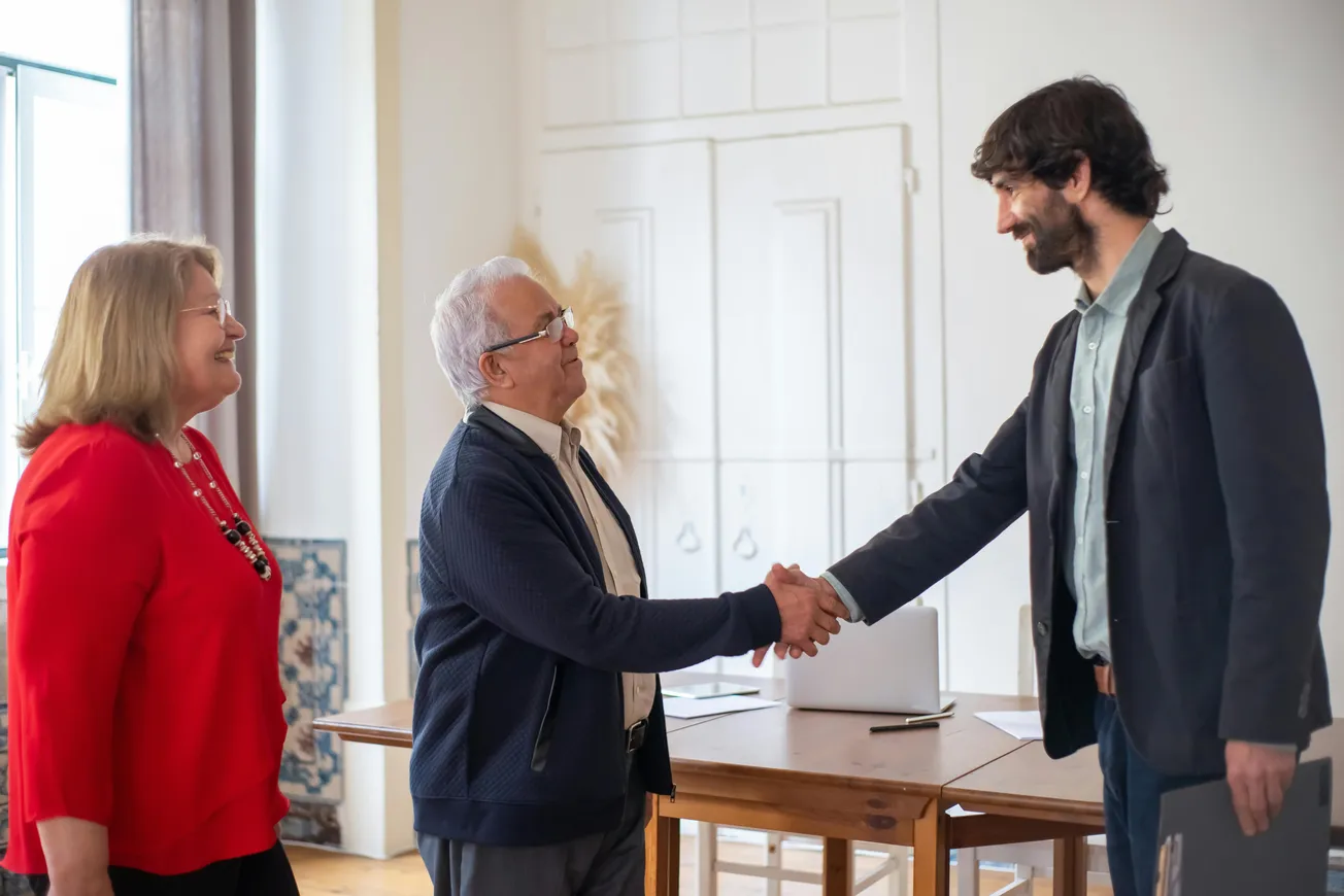 An elderly man and a younger man shake hands warmly in an office, while a woman in a red blouse smiles beside them, conveying a positive, professional atmosphere.