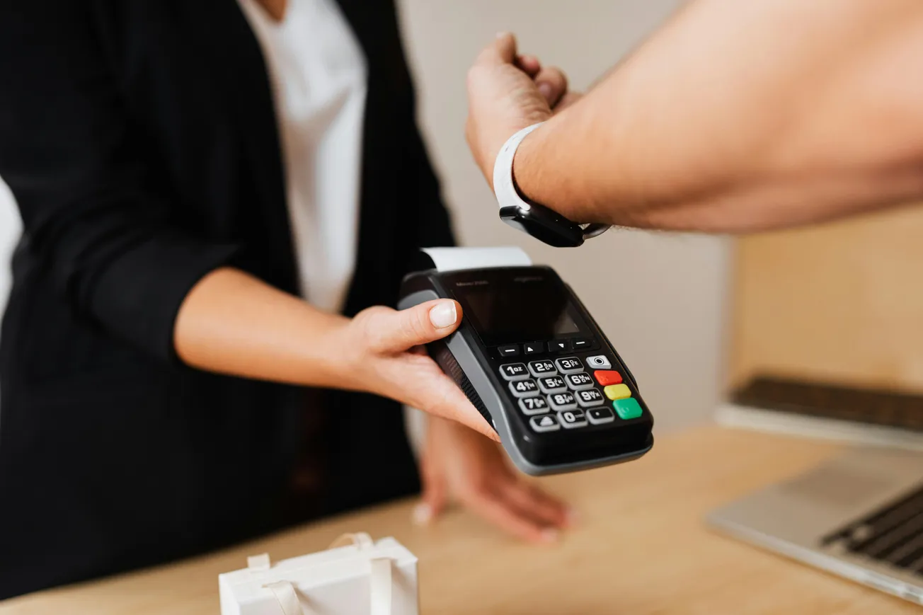 A person pays with a smart watch at a POS terminal held by a person in a black blazer, illustrating a modern, contactless transaction in a retail setting.