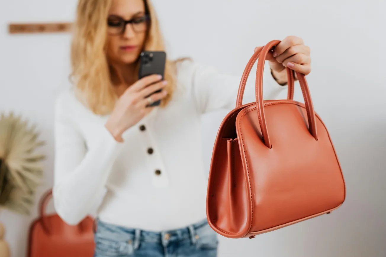 A woman in a white shirt and glasses takes a photo of a stylish, coral leather handbag. She holds a smartphone, capturing the bag’s elegant design.