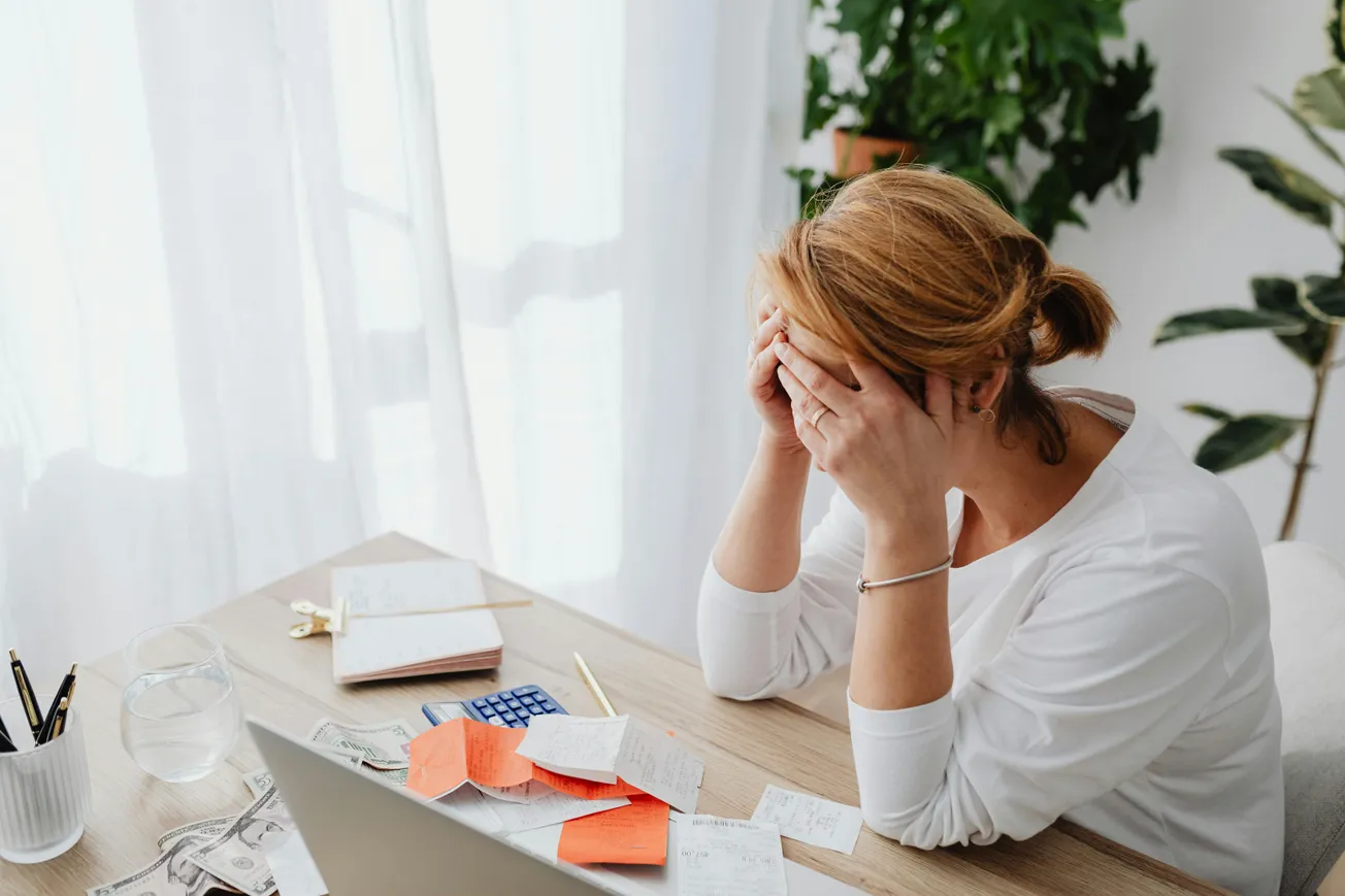 A woman sits at a cluttered desk with bills and notes, resting her head in her hands. The scene suggests stress amid paperwork and financial concerns.