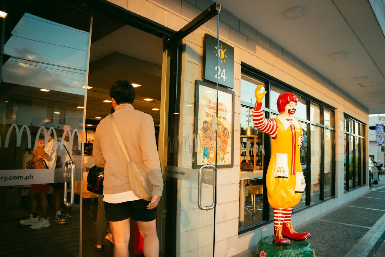 A person enters a brightly lit fast food restaurant. A Ronald McDonald statue waves outside. The scene conveys a welcoming, casual atmosphere.