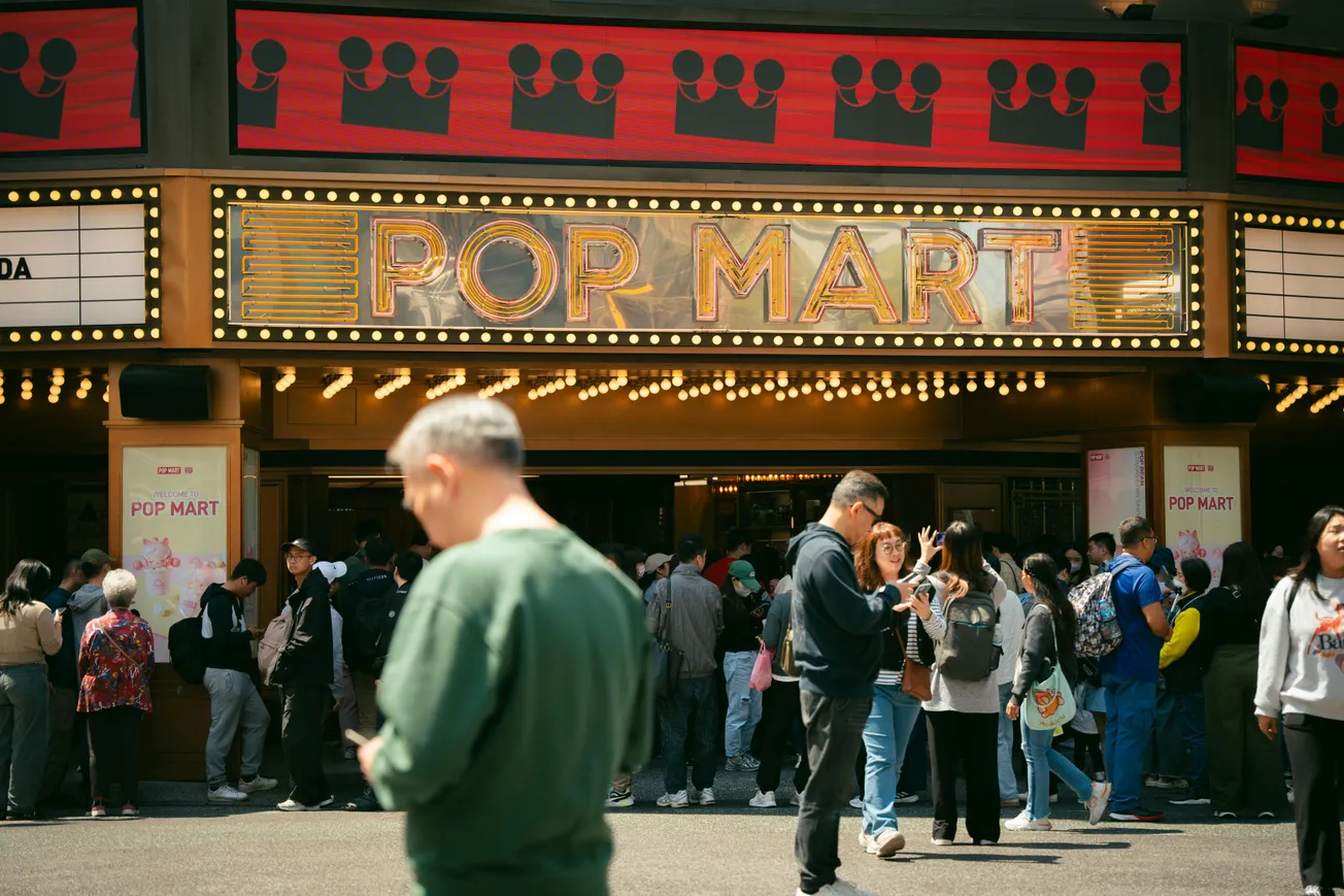 A crowd gathers outside a brightly lit theater with a marquee sign reading "Pop Mart." People chat and look at their phones, creating a lively, bustling atmosphere.