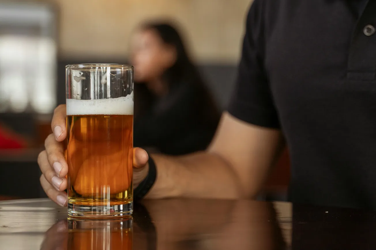 A person in a black shirt holds a glass of amber-colored beer with foam on top. The setting is a casual restaurant or bar, and a blurred figure is in the background.