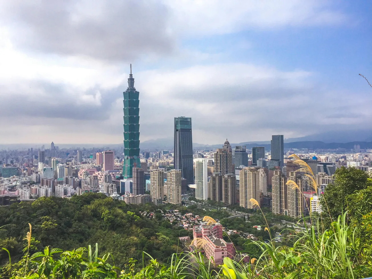 A vibrant cityscape of Taipei with Taipei 101 skyscraper standing out. Lush green foreground and a cloudy sky add contrast, creating a dynamic urban scene.