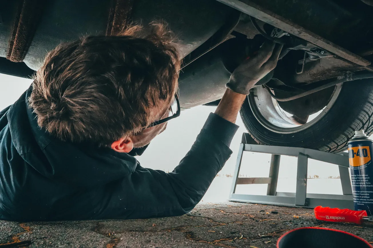 A person wearing glasses and gloves is working underneath a car on a lift. Nearby, there's a bright red tool and a can of WD-40, suggesting repair work.