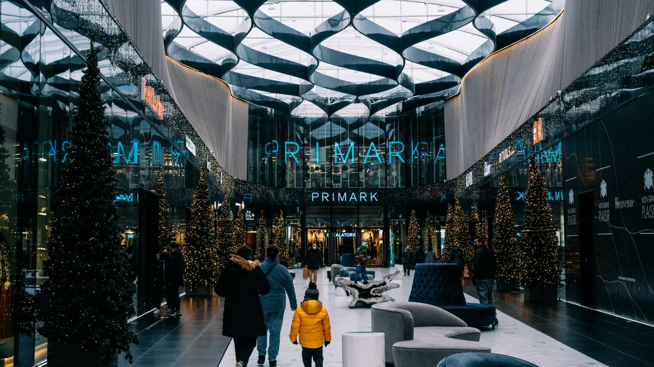 A modern shopping mall with a glass roof. People walk toward a store with a neon sign. Decorated Christmas trees and festive lights create a warm holiday atmosphere.