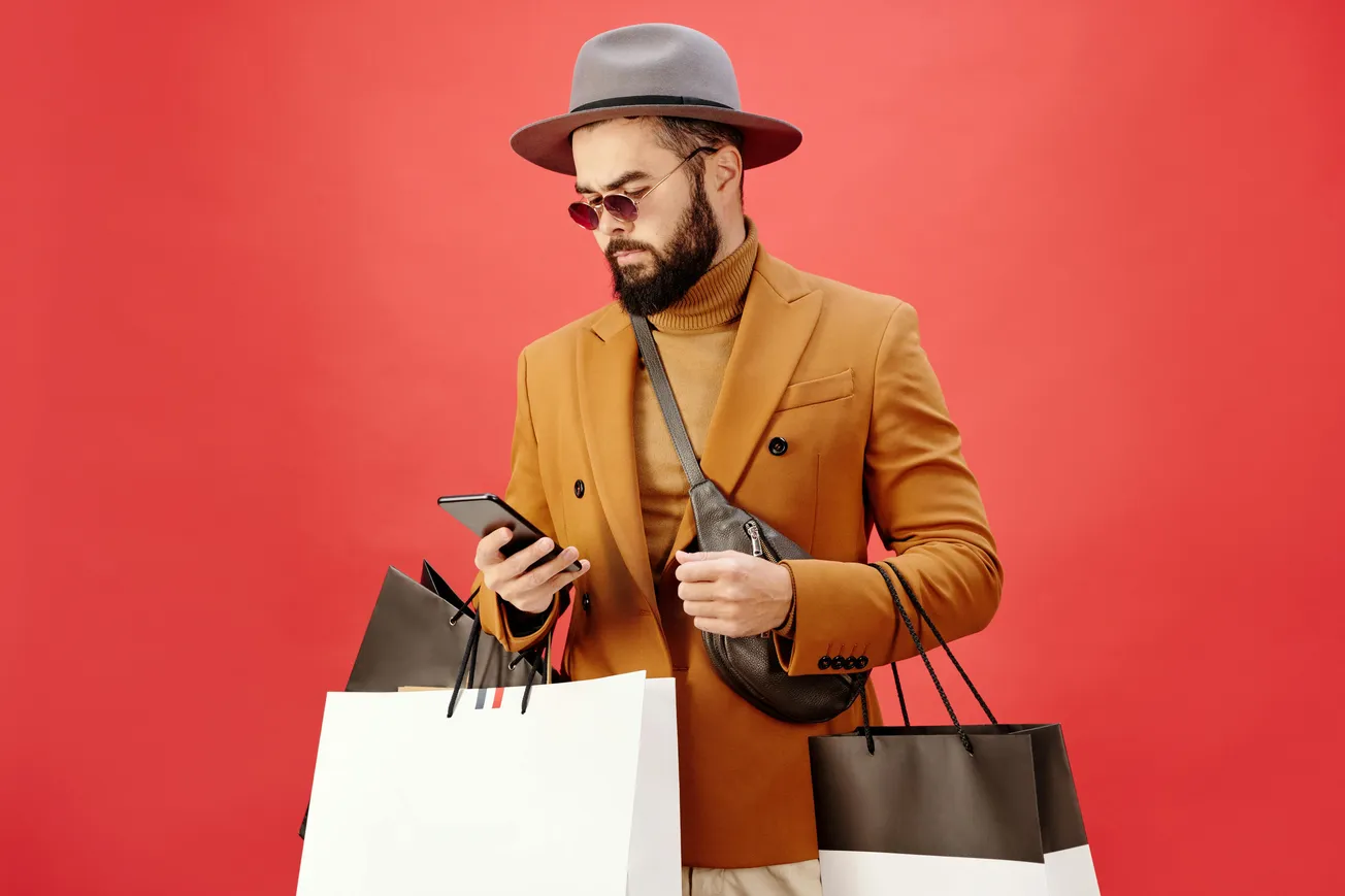 A stylish man in a brown blazer and hat holds shopping bags and looks at his phone against a red background, conveying a modern, fashionable vibe.