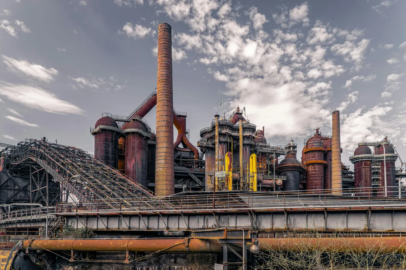 Alt text: A large, rusted industrial complex with towering chimneys and pipes set against a partly cloudy sky. The scene conveys an abandoned, historical atmosphere.