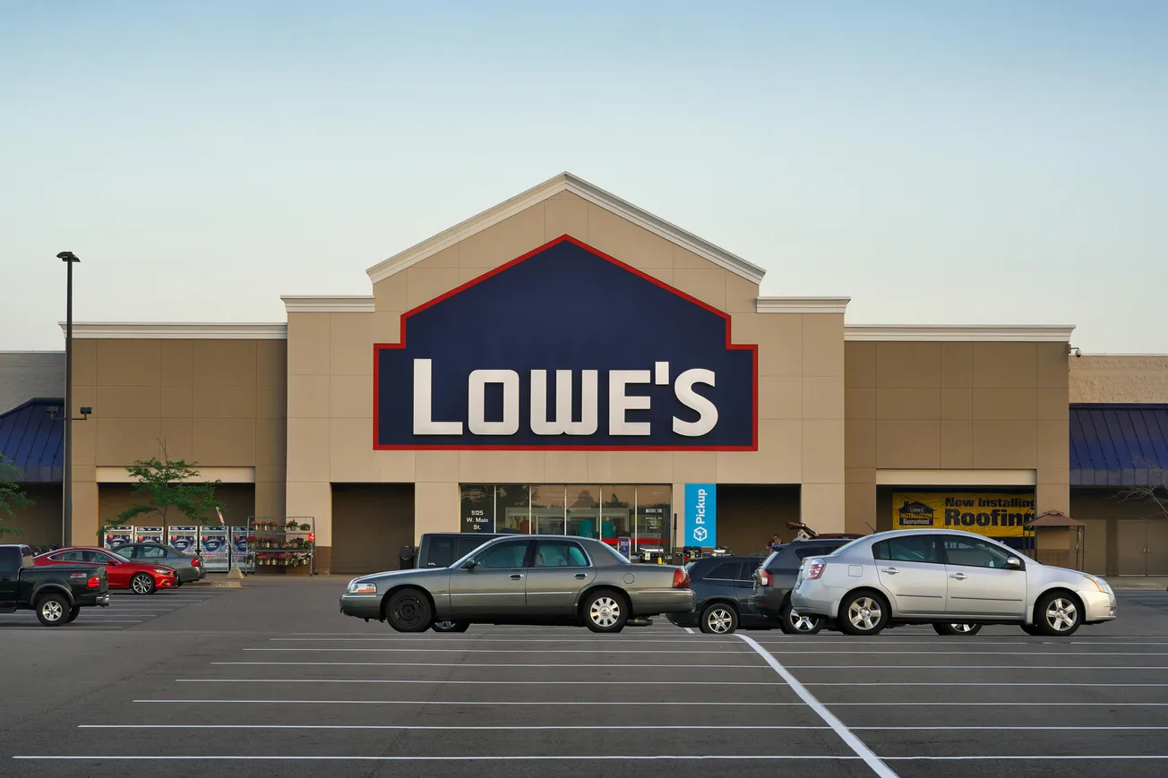 Storefront of a Lowe's home improvement store with a large blue and red sign. Several cars are parked in the lot in the early evening.