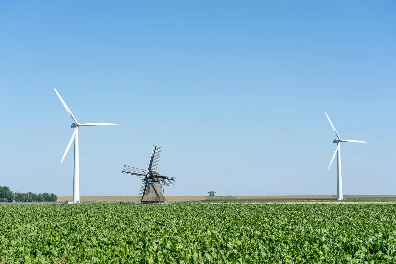 Two modern wind turbines and a traditional windmill in a green field under a clear blue sky, symbolizing the contrast between old and new energy sources.