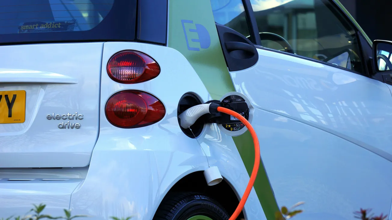 A white electric car is plugged into a charger. The charging cable is orange, and the car is labeled "electric drive." The scene conveys eco-friendliness.