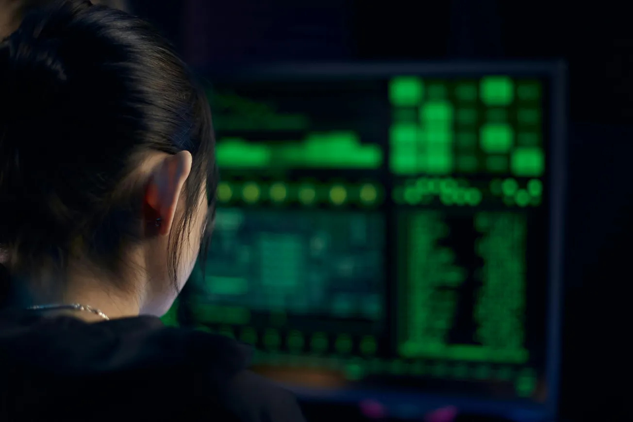 A person with dark hair is focusing on a computer screen displaying green text and graphics, suggesting a cybersecurity or programming setting.